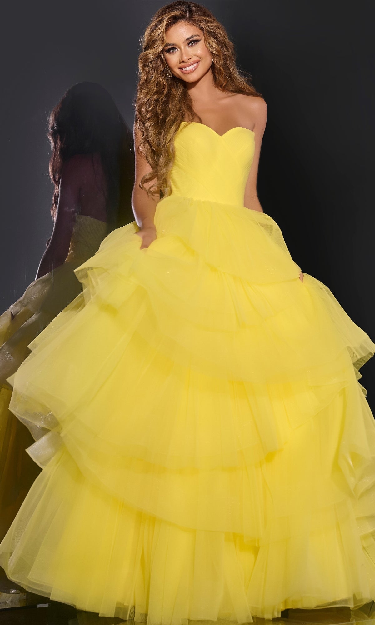 A woman smiles in front of a dark background wearing the Jovani 40394 Strapless Ruffled Prom Ball Gown in layered yellow, her reflection visible beside her.